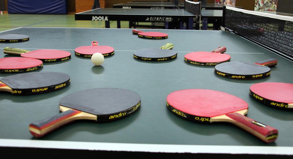 Mehrere Tischtennisschläger in verschiedenen Farben und ein Tischtennisball liegen auf einer grünen Tischtennisplatte.